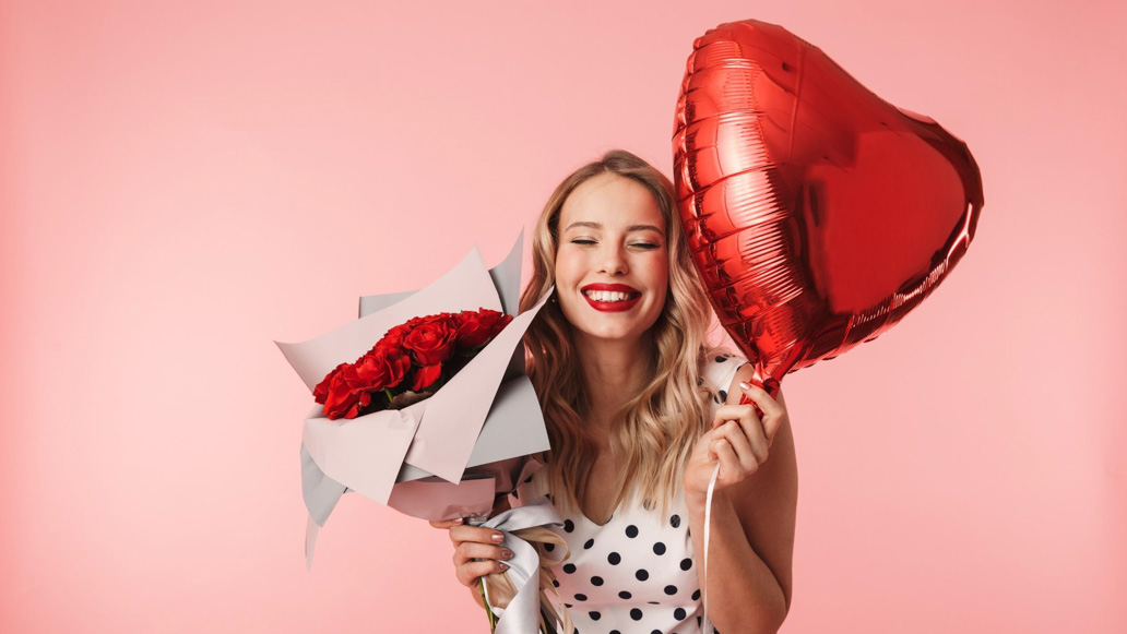 Νεαρή κοπέλα χαμογελά με μπαλόνι σε σχήμα καρδιά και κόκκινα τριαντάφυλλα στο χέρι