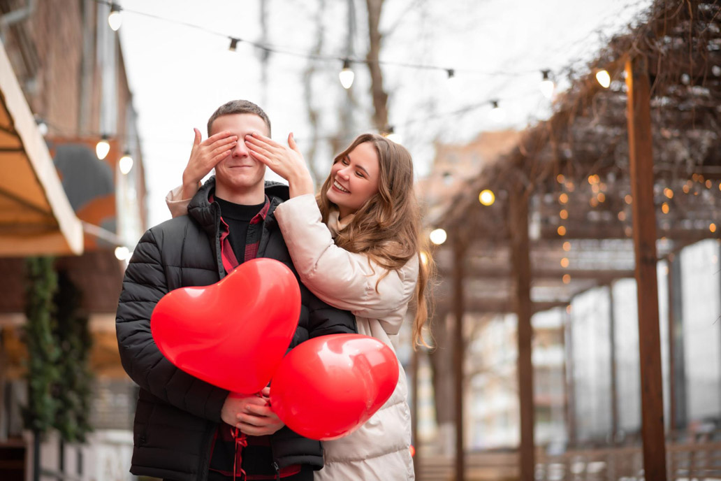Ζευγάρι ερωτευμένων έχει βγει ραντεβού, με νεαρό άντρα να κρατά μπαλόνια και τη γυναίκα να του κλείνει τα μάτια.