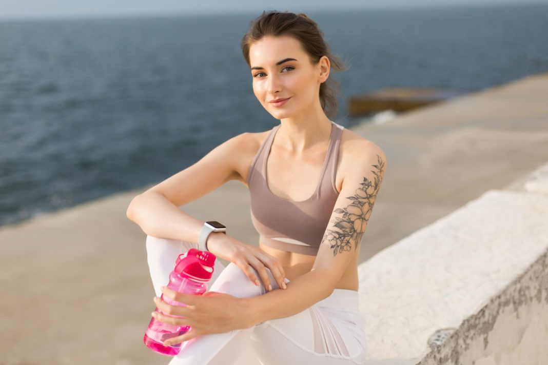 Νεαρή χαμογελαστή γυναίκα με αθλητική ενδυμασία και ένα ροζ αθλητικό μπουκάλι κοιτάζει την κάμερα με φόντο τη θάλασσα.