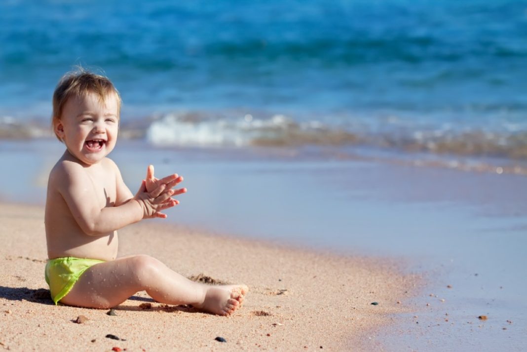 Χαρούμενο μωρό αγόρι κάθεται στην παραλία δίπλα στο κύμα.